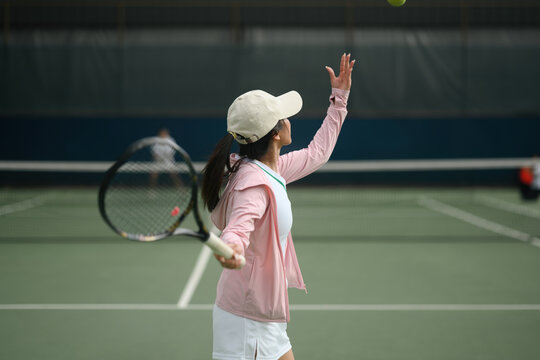 Young Asian Woman Serving Tennis Ball on Outdoor Court in Pink Sportswear and White Cap