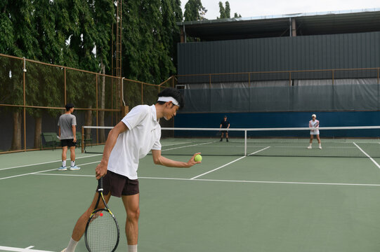 Young Asian Man Preparing to Serve on Outdoor Tennis Court with Players in Background