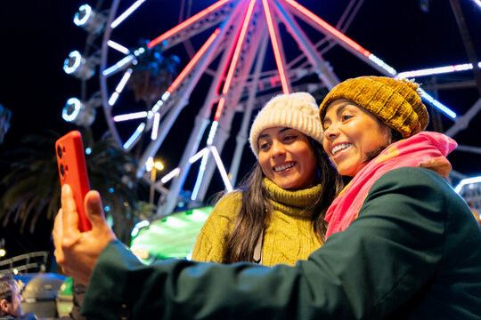 Two happy women enjoying night out at amusement park taking selfie with illuminated Ferris wheel in background