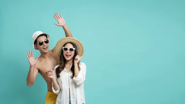 Happy young couple in summer clothes waving hands on isolated teal background