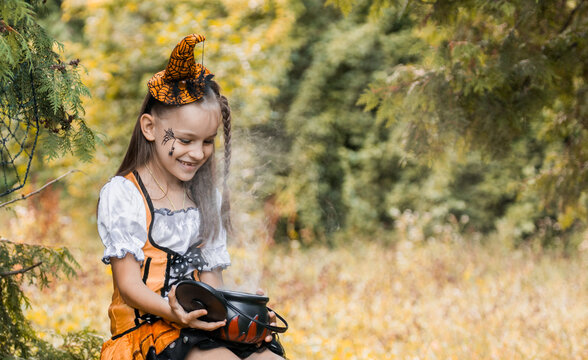 Girl in a Costume Sits in a Field During Autumn, Surrounded by Colorful Trees and Halloween Decorations in the Month of October