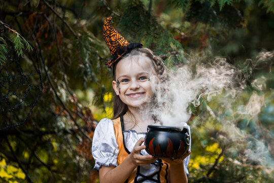 Cute Girl in Halloween Costume Holding a Cauldron With Smoke in a Garden With Autumn Leaves and Spiderwebs Around Her