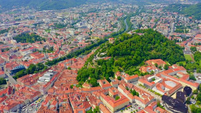 Graz, Austria. Aerial panorama over the historic city center and Mount Schlossberg, dense terracotta rooftops and the winding Mur River under clear summer light. Drone footage, Departure of the camer