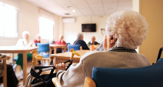 Back view of senior woman with white hair sitting beside a walker in a nursing home, with a group of elderly people socializing in the background. Senior care, assisted living and retirement concept