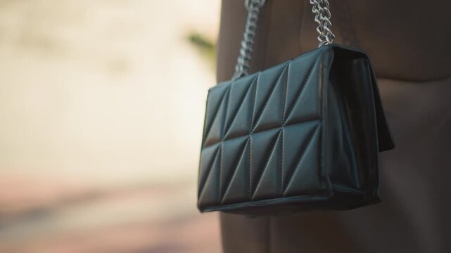 Close up of black quilted leather handbag with silver chain strap hanging from arm. bag features geometric stitching details and soft lighting highlights set against blurred background
