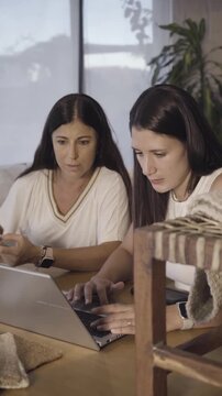 Two women lean in closely at a wooden table, focused on inspecting woven carpet samples together in soft natural light. Their