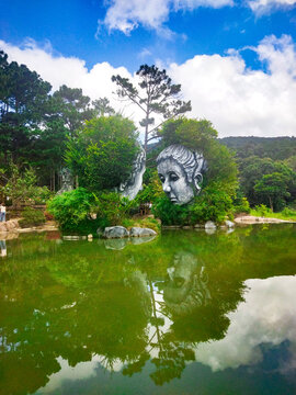 Giant sculpted faces rise beside a lake at God Valley near Da Lat, Vietnam. Reflections and pine forest frame the scene. Suitable for travel, tourism, fantasy parks and destination articles