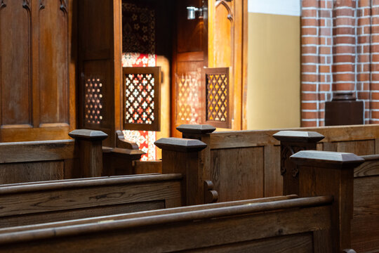 Traditional wooden church pews and carved wooden confessional booth illuminated by warm sunlight, historic church interior with brick wall