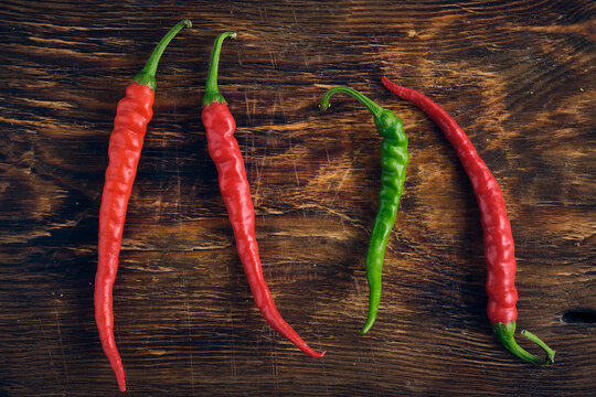 Four long spicy chili peppers three red and one green laid out on dark textured wooden background.