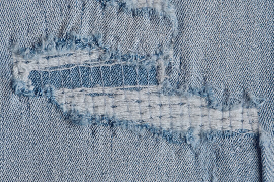 Detailed view ripped blue denim fabric with exposed white threads and distressed texture.