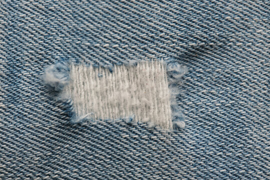 Close up texture light blue denim jean fabric featuring rectangular frayed tear with exposed threads.
