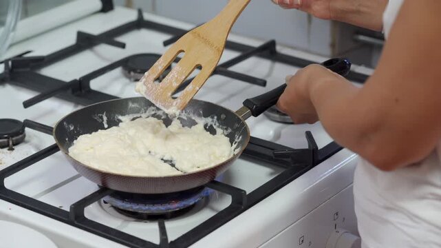 A careful cook gently scrambles a creamy egg mixture in a hot pan. She prepares a comforting homemade breakfast on the kitchen stove with patient dedication.