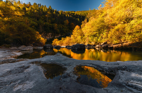 A wide autumn landscape of golden and orange forest hillsides reflected in a still river pool, with rocky foreground and a bright blue sky.