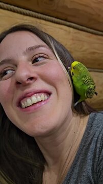Woman smiles warmly with a small green parakeet perched gently on her shoulder. Bird rests close to her cheek, eyes soft, feathers bright green. Wooden wall background adds cozy. Parakeet appears calm