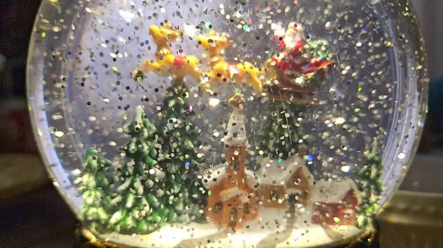 Snow globe with festive Christmas scene. Small reindeer and Santa Claus float inside. Trees and a cozy house sit beneath falling glitter. Snowflakes swirl around the miniature scene