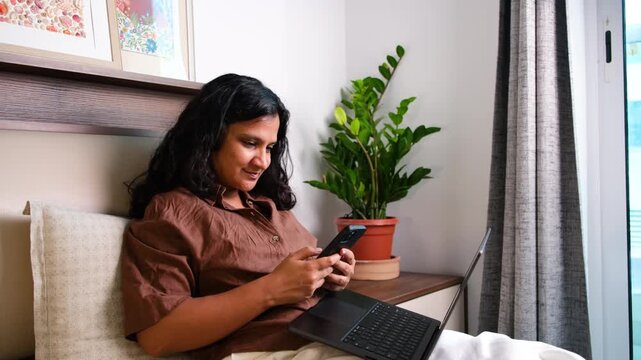 Woman distracted by smartphone while working on laptop at home, social media scrolling, multitasking, procrastination and remote work productivity concept