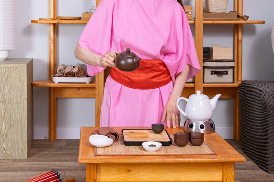 Woman wearing pink kimono performing traditional tea ceremony. Tea master pouring beverage from clay teapot into cup. Zen culture, mindfulness, oriental lifestyle and wellness concept