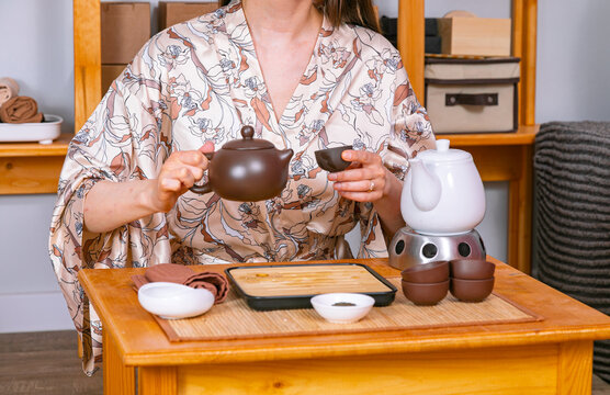 Woman wearing floral robe performing traditional tea ceremony. Tea master pouring beverage from clay teapot into cup. Ritual of preparing organic green tea drink in calm home interior