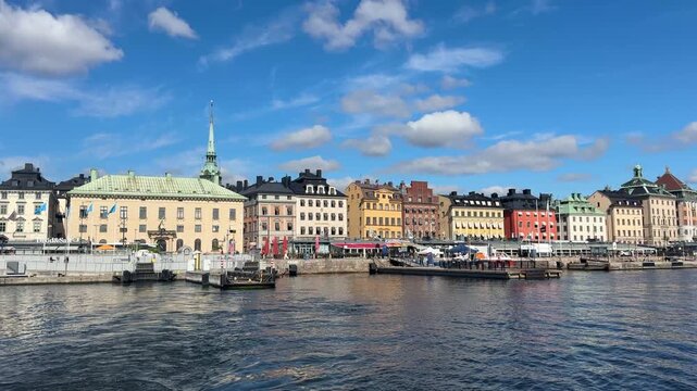 Wide scenic boat point-of-view tracking shot past the iconic colorful medieval buildings, church spires, and piers of Skeppsbron in Gamla Stan, Stockholm under a bright blue summer sky.