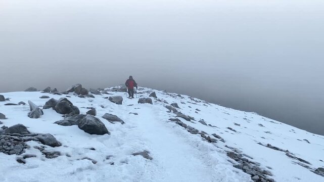 Mountaineer trekking in storm wind in wide view on Mount Ararat, crossing a snowy rocky slope in dense fog for extreme weather, alpine safety, expedition, endurance, survival, and travel use