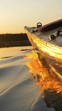 Close up view showing the bow of a kayak sailing on a tranquil lake during a vivid golden sunset, with the brilliant light reflecting and shimmering across the rippling water surface