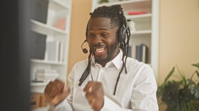 Man with headset at computer desk, fists raised and smiling, eyes closed and clapping hands during a virtual win in studio; workplace joy.