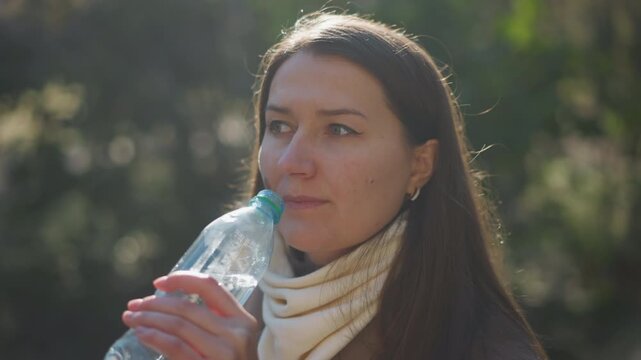 Close up of woman drinking water outdoors in soft natural light. Woman holds and drinks from clear plastic bottle wearing white scarf with long brown hair visible. background is blurred