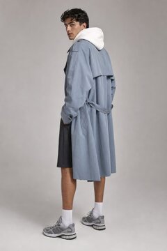 Young man in stylish light blue oversized trench coat with white hoodie lining, wearing white socks and athletic shoes, casual urban streetwear fashion look, minimalist portrait against neutral