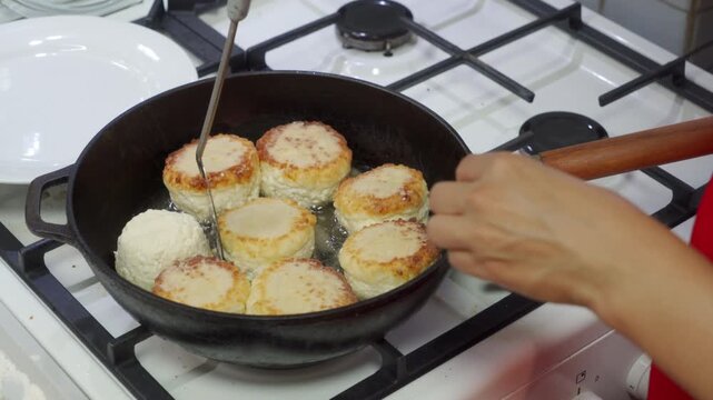 An attentive cook skillfully flips golden cottage cheese pancakes in a sizzling frying pan. She works happily at a clean gas stove inside a warm, busy kitchen.