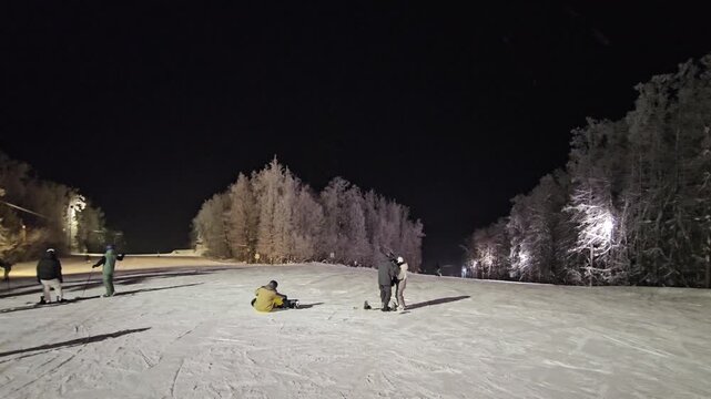 Night ski slope bathed in artificial light. Snowy trees glow under distant illumination. Skiers pause near a seated figure on the slope. Scene captures quiet winter recreation after dark