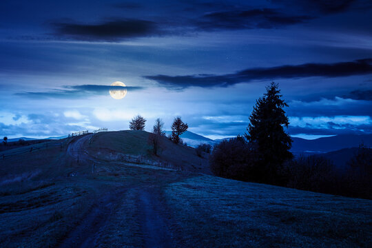 winding rural dirt path through hill at night. rolling countryside of ukraine in fall color. cloudy sky in full moon light. carpathian mountains in autumn