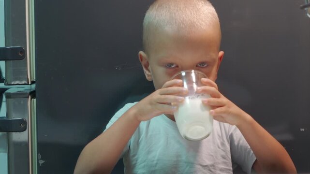 A focused little boy drinks fresh milk from a glass in a kitchen. He looks directly forward with serious intensity, finding peaceful satisfaction in his nutritious beverage.