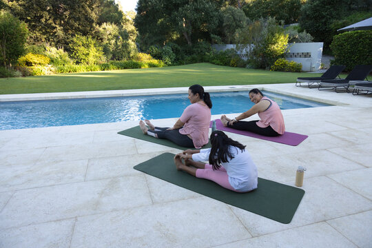 Diverse female family stretching on yoga mats beside pool on tiled patio with water bottle