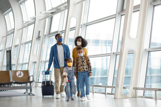 A Black family with two children wearing face masks and carrying luggage walks through a modern airport terminal