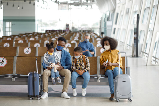 A diverse family with young children sits in an airport waiting area, wearing protective face masks and observing social distancing protocols while traveling