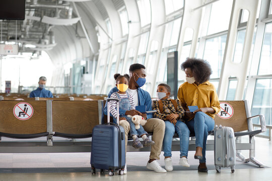 A diverse family with children sits in an airport departure lounge wearing protective face masks and holding passports while waiting for their plane