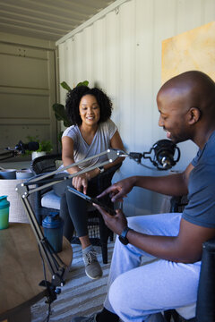 Diverse podcast hosts leaning forward, pointing at tablet, recording with mic booms in metal studio
