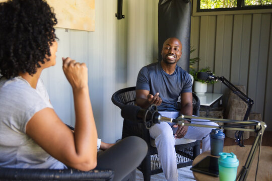 Diverse cohosts speaking and smiling in sportswear in home studio with mic arms, tablet, shakers
