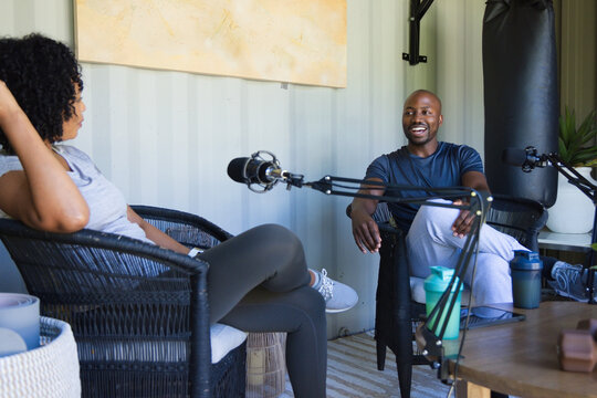Diverse cohosts wearing activewear, sitting opposite, recording chat on patio with mics, punch bag