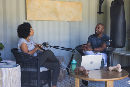 Diverse co-hosts wearing casual athleisure recording podcast in studio with mics laptop dumbbells