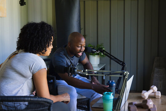 Diverse co-hosts leaning forward recording podcast at home studio with laptop microphones dumbbells