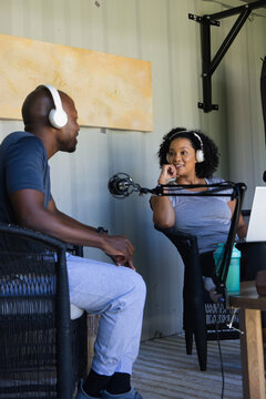 Diverse coworkers wearing white over-ear headphones recording podcast at studio with mic, laptop