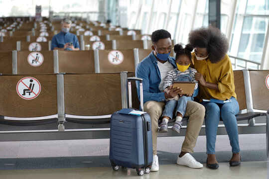 A Black family with a young child sits together in an airport waiting area, all wearing face masks and looking at a tablet, with social distancing markers visible on the seats