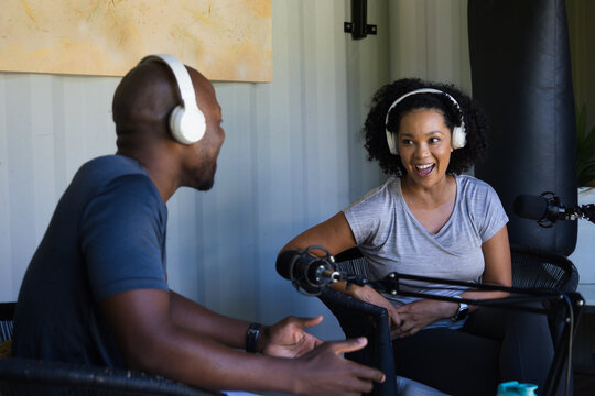 Diverse co-hosts recording lively podcast in small studio using white headphones and boom mics