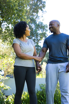 Couple standing holding hands smiling talking in garden wearing athletic clothing by low white wall