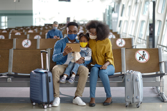 A family with two children wearing face masks sits in an airport waiting area with their luggage and a tablet device