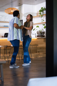 Diverse couple holding hands and swaying in modern kitchen near wooden island and coffee machine