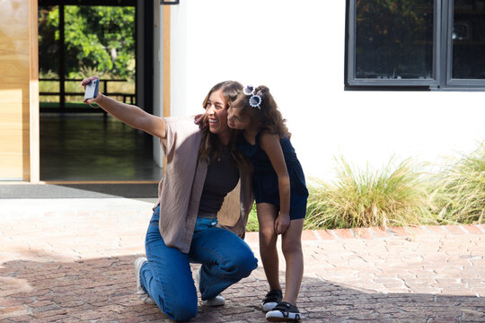 Female caregiver kneeling and holding phone, girl leaning in and smiling on brick patio