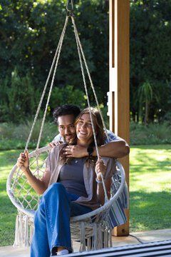 Diverse couple sitting on hanging macrame swing chair at backyard porch holding braided ropes