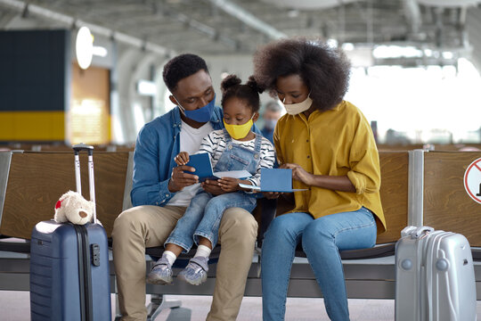 A mother, father, and young daughter sit together in an airport terminal, all wearing protective face coverings while reviewing their travel documents